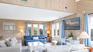 Un salon spacieux avec un canapé bleu, un canapé blanc et une table basse en verre. De grandes fenêtres sont ornées de rideaux bleus, un tableau est accroché au mur et une petite table avec une lampe se trouve dans un coin.