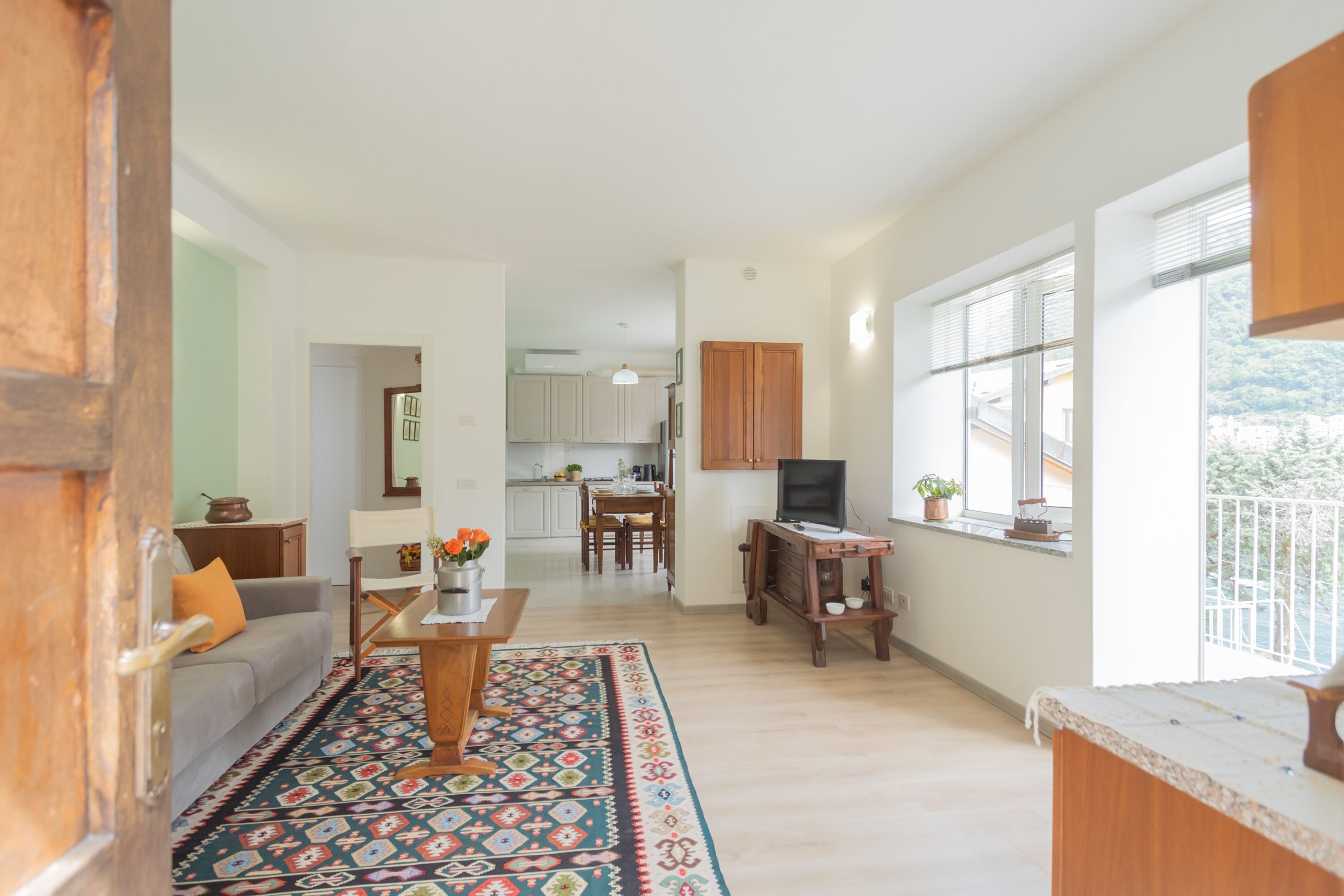 Sala de estar com sofá, mesa de centro e tapete estampado. Há uma TV sobre um suporte de madeira, uma área de jantar com cadeiras e uma cozinha ao fundo.