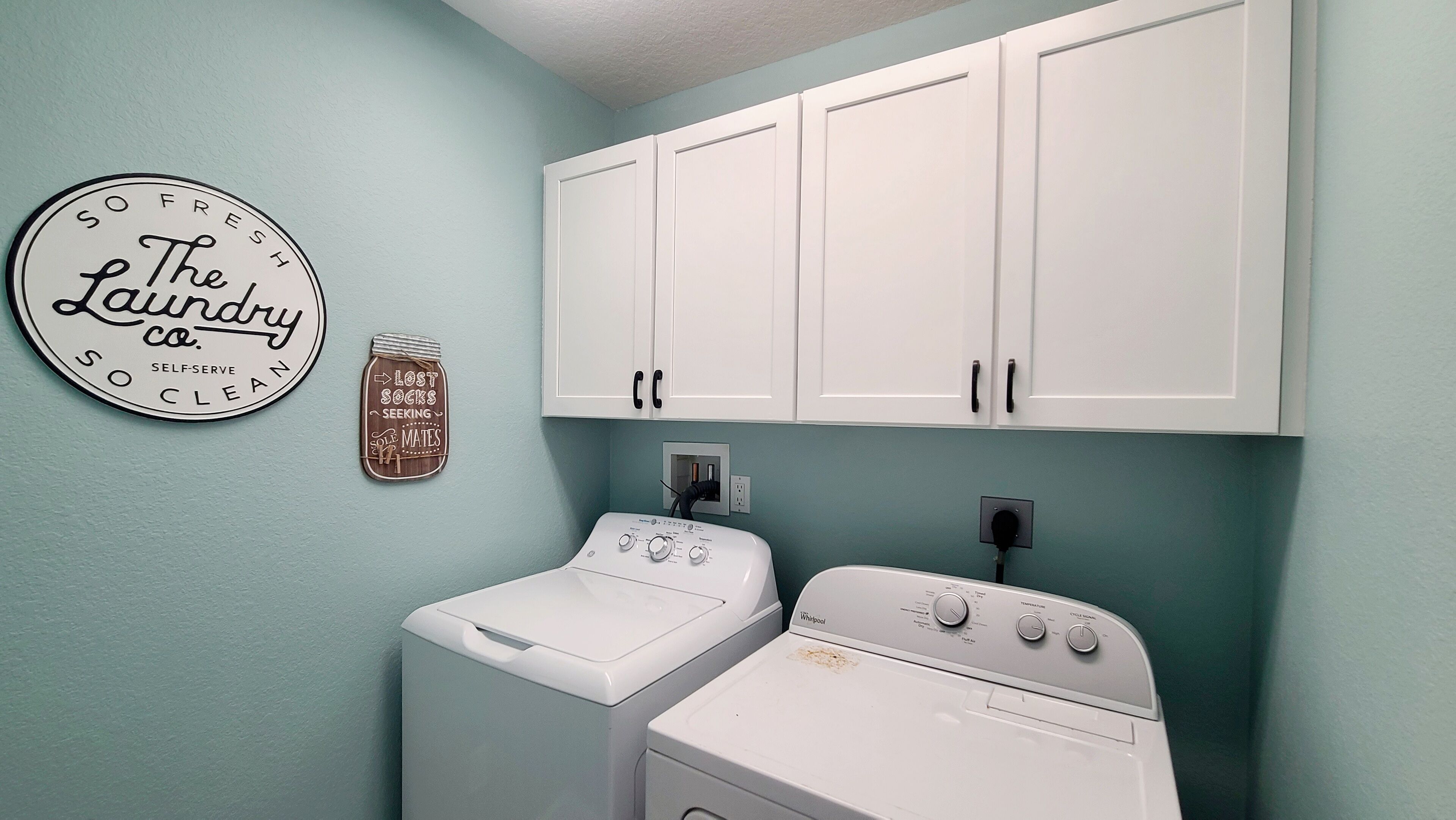 A laundry room with a wall-mounted circular sign, a jar-shaped sign, and a front-loading washer and dryer.