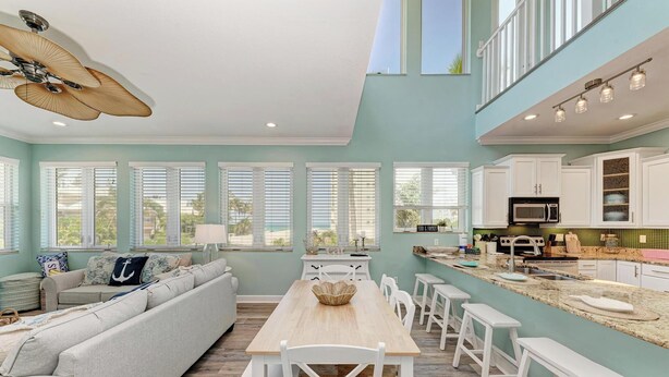 A spacious living room with a large sofa, a wooden dining table, and white bar stools. There is a kitchen area with white cabinets and a microwave. - Coquina Sands - Pool Home with Beach Views - Powered by Island Real Estate (Holmes Beach)