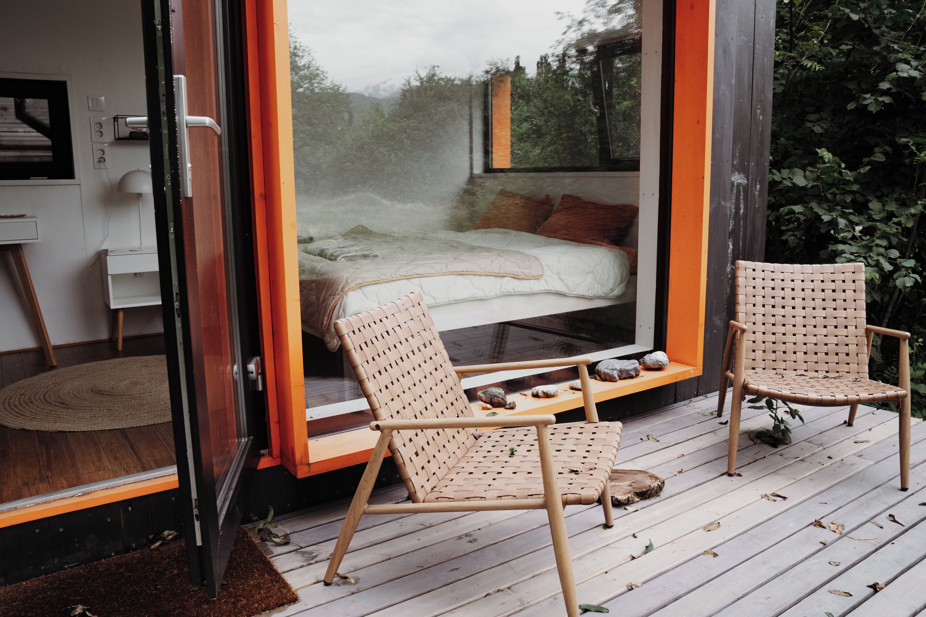 A modern bedroom with a large window, a bed, and two wicker chairs on a wooden deck.