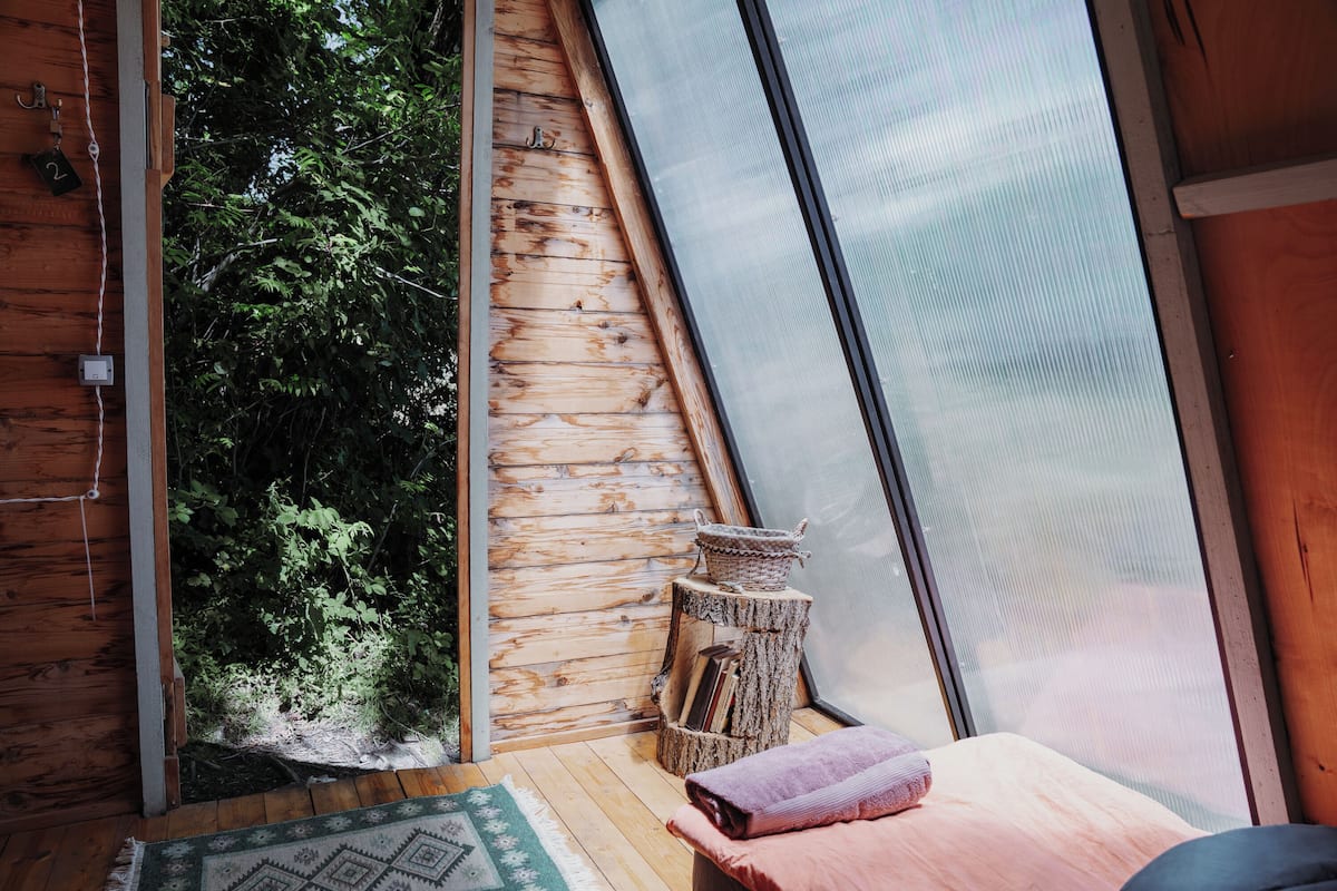 Une pièce chaleureuse avec un plancher en bois, un lit recouvert d’une couverture rose, un panier tressé posé sur un tronc d’arbre, et une fenêtre donnant sur la verdure.