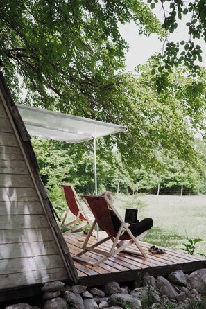 A wooden deck with two chairs under a canopy, surrounded by greenery.