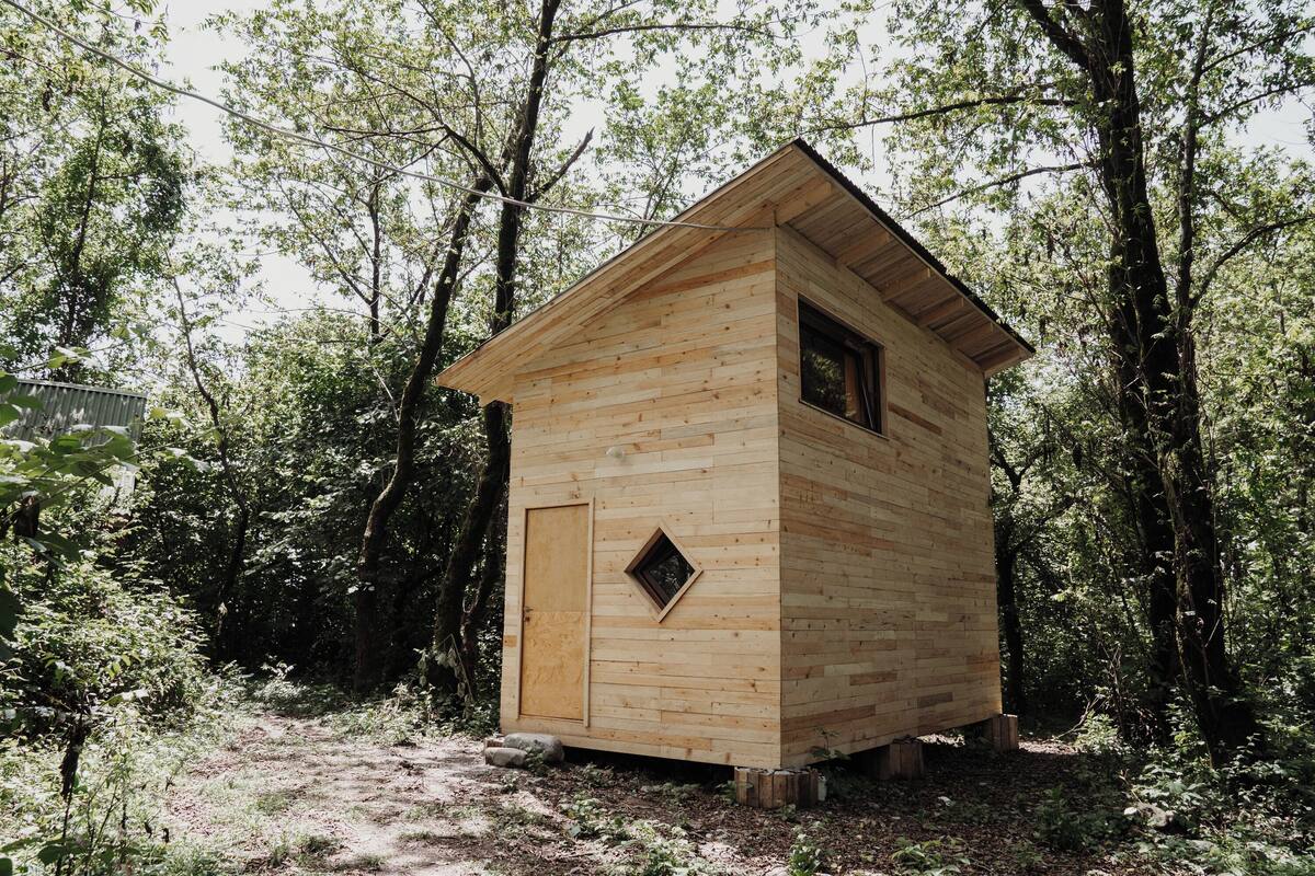 Une cabane en bois avec un toit à deux versants, une petite fenêtre et une porte, entourée d’une forêt dense.