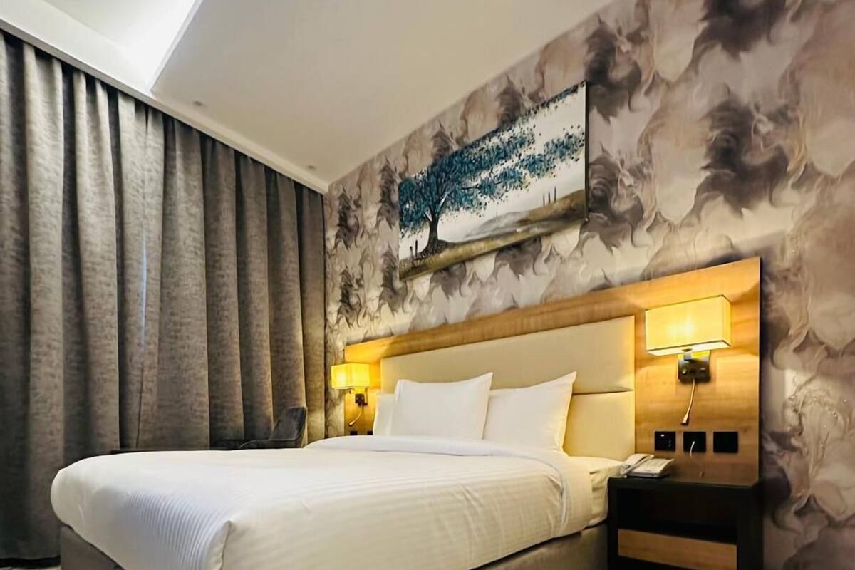 A hotel room with a large bed, a headboard with a built-in lamp, a nightstand with a phone, and a wall with a tree mural.
