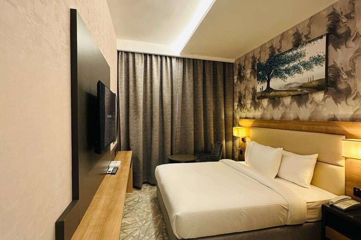A hotel room with a bed, a TV, a desk, and a painting on the wall.