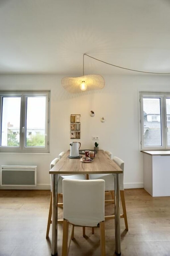 A dining area with a wooden table and white chairs, a hanging light fixture, and a window with a view.