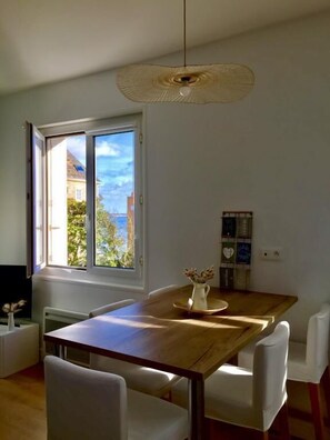 Un coin repas avec une table en bois et des chaises blanches, un luminaire suspendu et une fenêtre donnant sur des bâtiments et des arbres.