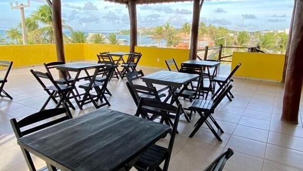 An outdoor patio with tables and chairs, a thatched roof, and a view of the ocean. - Pousada A Praia (Parnamirim)