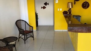 A room with a yellow accent wall, a white door, and a tiled floor. There are two wicker chairs and a small table. A lamp and decorative plates are on the wall. - Pousada A Praia (Parnamirim)
