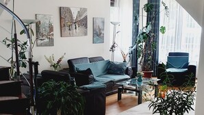 Ein geräumiges Wohnzimmer mit einer schwarzen Couch, einem Couchtisch und mehreren Blumentöpfen. Eine Treppe führt zu einer oberen Ebene, und der Raum hat einen Fliesenboden mit einem geometrischen Muster.