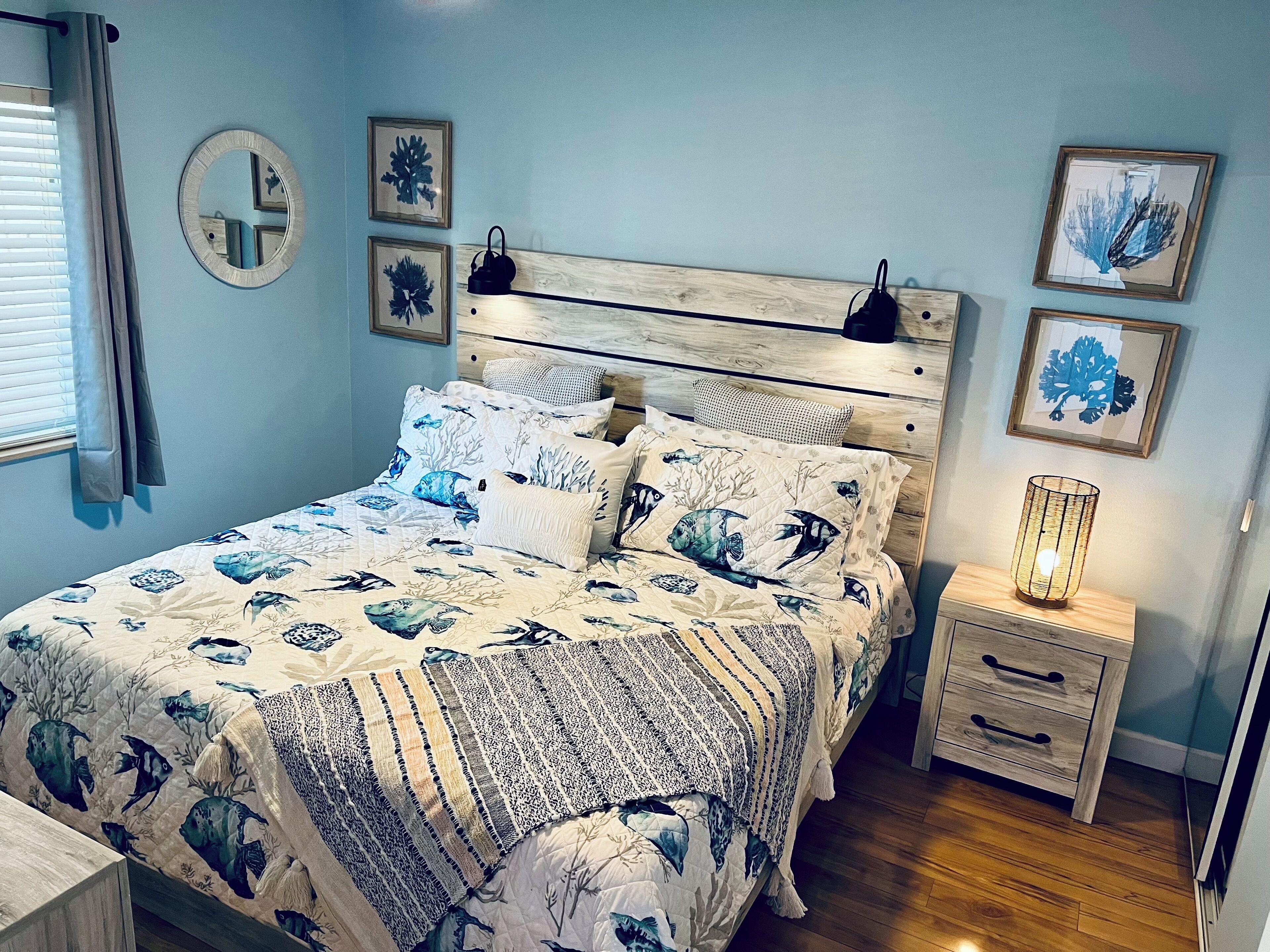 Un dormitorio con una cama que presenta una colcha de motivos marinos en azul y blanco, un espejo redondo y varias impresiones botánicas enmarcadas en la pared.