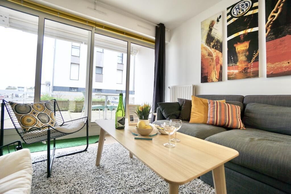 A modern living room with a sofa, a coffee table, and a chair. There are paintings on the wall and a window with a view of buildings outside.