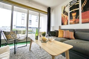 A modern living room with a sofa, a coffee table, and a chair. There are paintings on the wall and a window with a view of buildings outside.