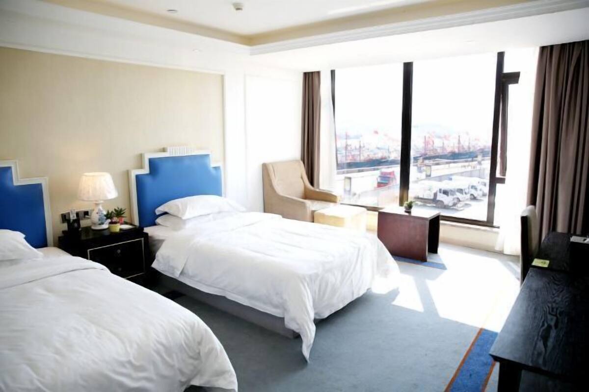 A hotel room with two beds, a desk, a chair, a window with a view, and a lamp.