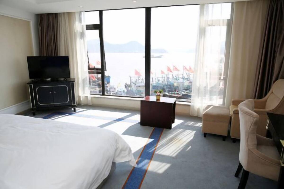 A hotel room with a bed, a TV, a small table, a chair, and a view of the sea.
