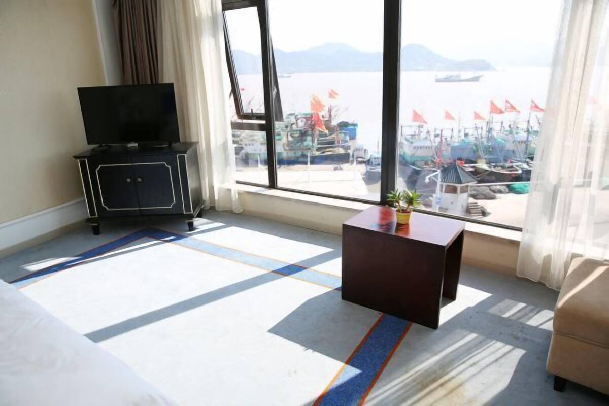 A hotel room with a bed, a TV on a cabinet, a small table with a plant, and a view of a harbor with boats and flags.