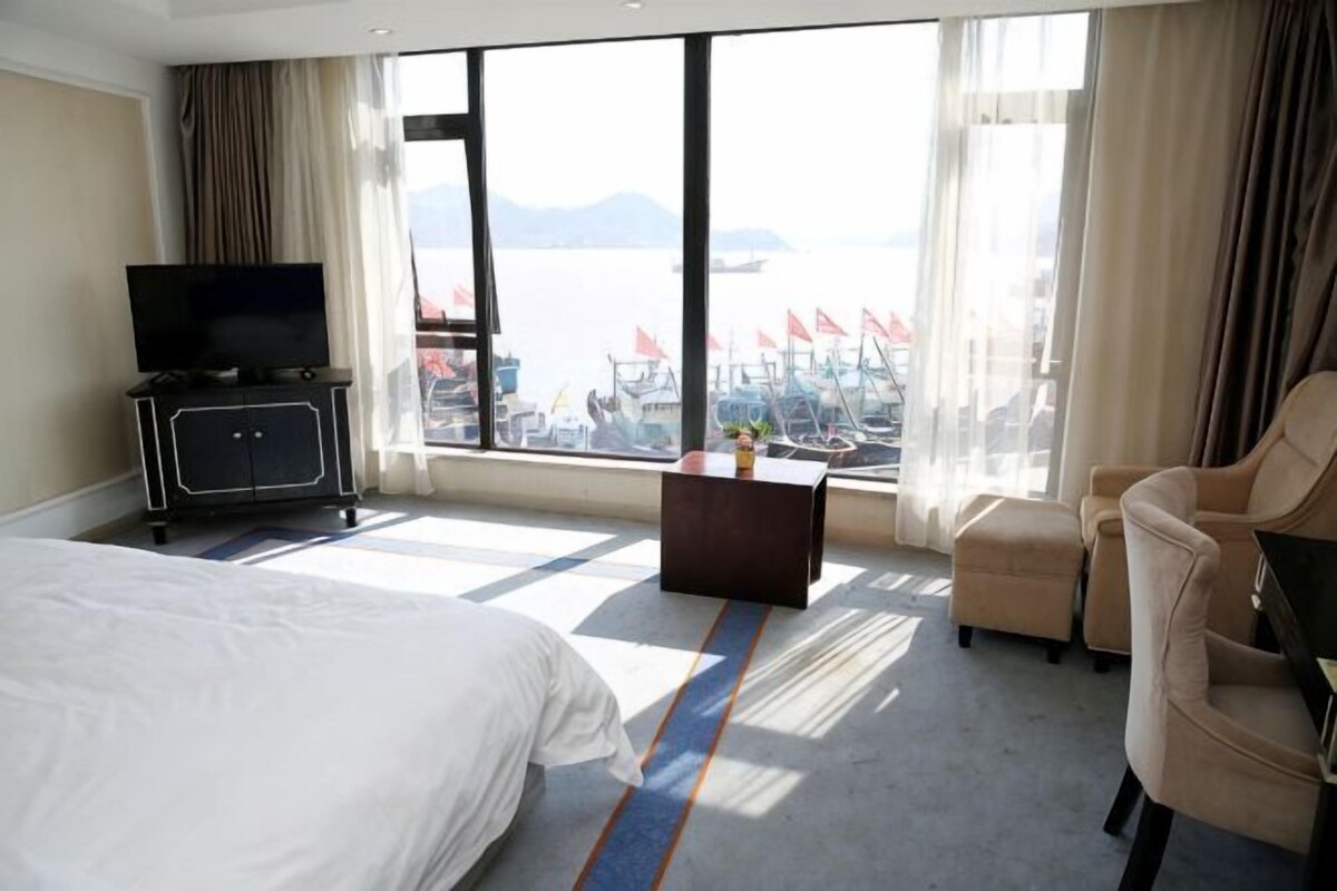 Een hotelkamer met een bed, een televisie op een standaard, een bureau, een stoel en een groot raam met uitzicht op boten en water.