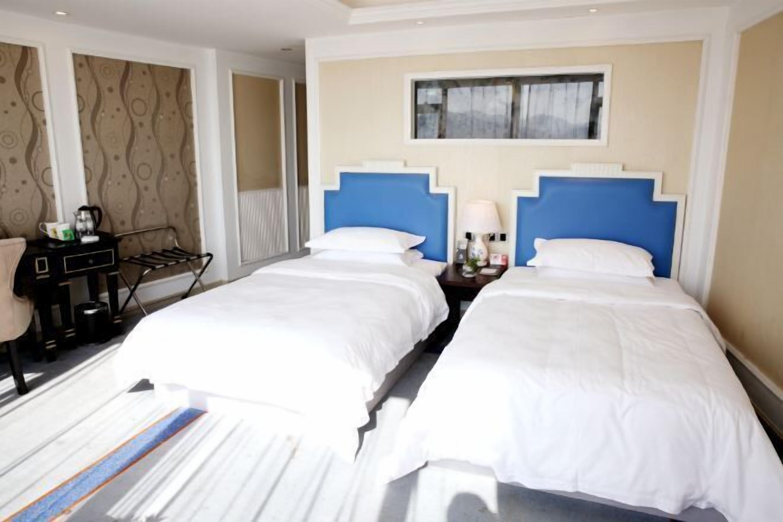 A hotel room with two beds, a desk with a chair, a small table with a lamp, and a window with a view.