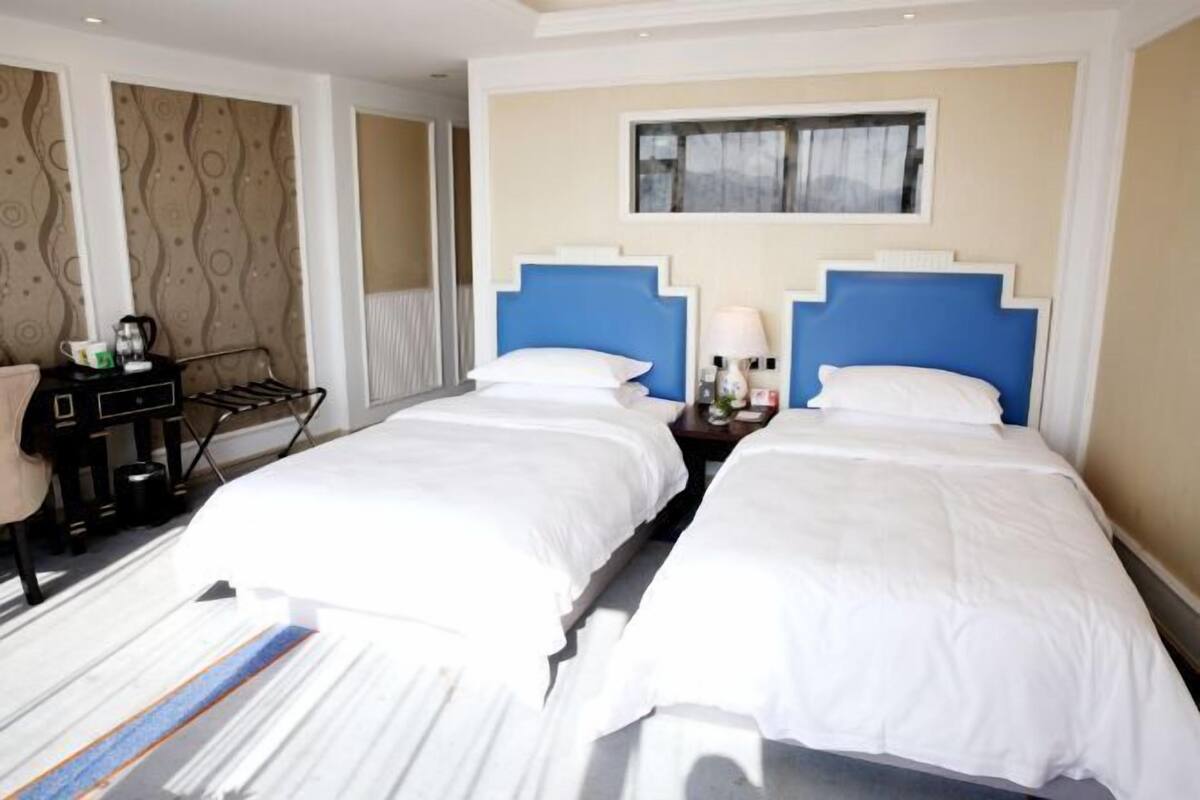 A hotel room with two beds, a desk with a chair, a small table with a lamp, and a window with a view.