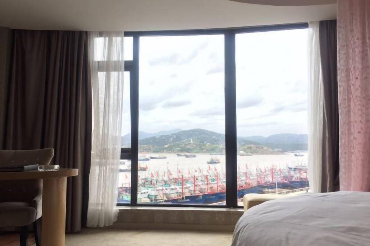 A hotel room with a large window, a bed, a chair, and a small table.