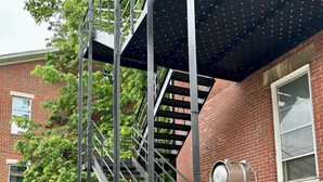 Una escalera metálica con techo metálico, que conduce a un edificio de ladrillo con ventanas y un carretel de manguera contra incendios.