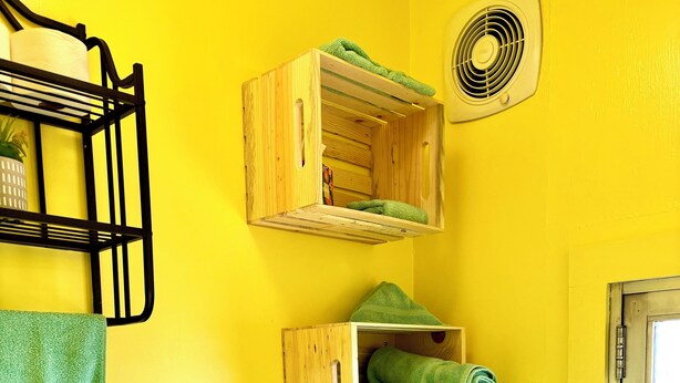 Un baño con paredes amarillas, un estante de madera con toallas, un inodoro, una planta y una ventana.