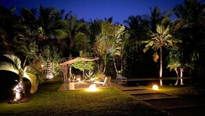 Un espacio exterior bien iluminado con una zona de estar cubierta, piscina y exuberante vegetación tropical.