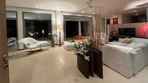 Habitación de hotel moderna con una cama grande, un rincón de descanso con una silla blanca, televisión y un balcón con vistas al cielo nocturno.