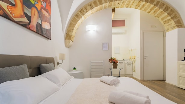 Une chambre d’hôtel avec un grand lit, une tête de lit, un tableau au mur, une petite table sur laquelle est posé un vase de fleurs, et une porte en arc donnant accès à la salle de bain.