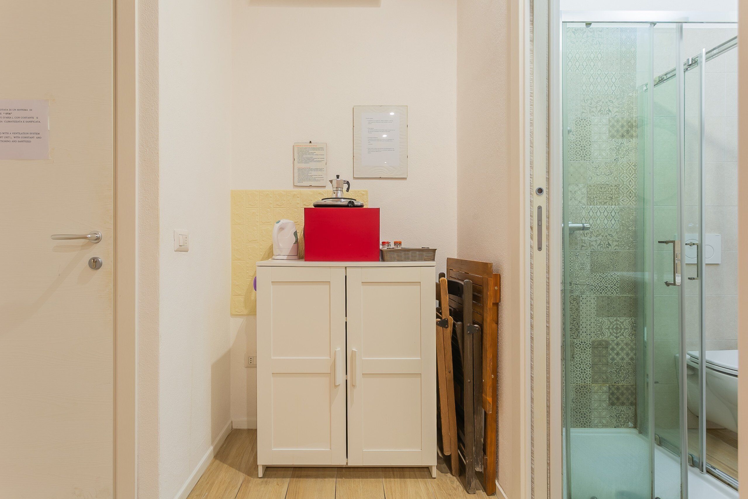 A small, compact bathroom with a shower, a white cabinet with a red appliance, and a wooden chair.