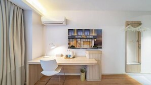 Ett modernt rum med ett skrivbord i trä, en vit stol, ett litet bord med en lampa och en väggmonterad luftkonditioneringsanläggning.