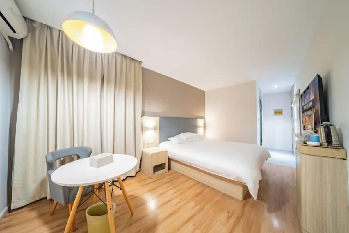 A hotel room with a bed, a desk, a chair, a small table, a nightstand, and a TV.