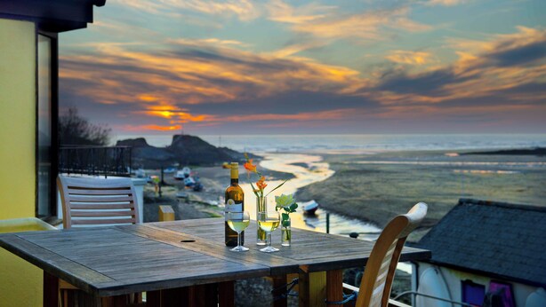 A table set with a bottle of wine, glasses, and a flower arrangement, overlooking a beach with a sunset in the background.
