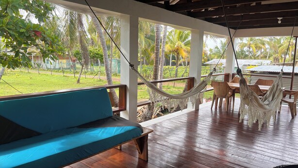 A hammock chair and a blue sofa on a wooden deck with a view of a grassy area and palm trees.