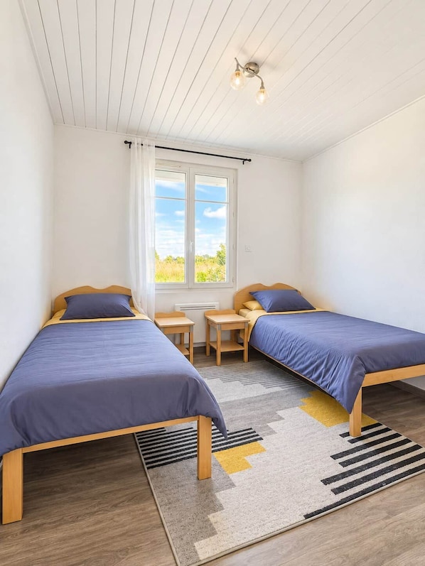 Quarto com duas camas de solteiro, janela com cortinas, uma mesinha e luminária de teto.