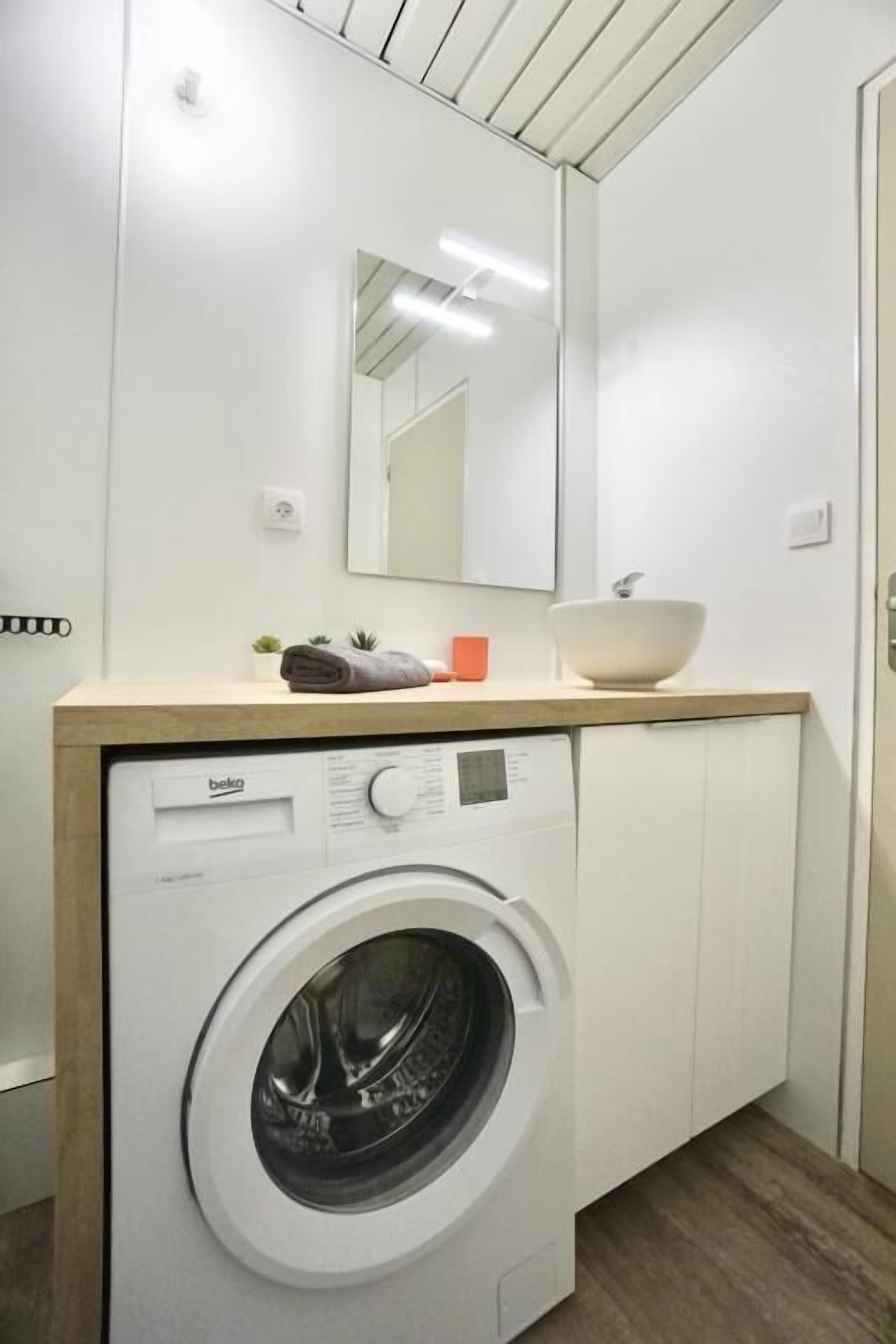 A compact laundry room with a front-loading washing machine, a sink, and a mirror above.