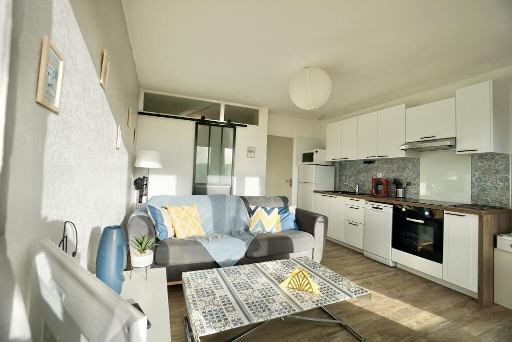 A modern living room with a sofa, a coffee table, and a kitchen area with white cabinets and appliances.