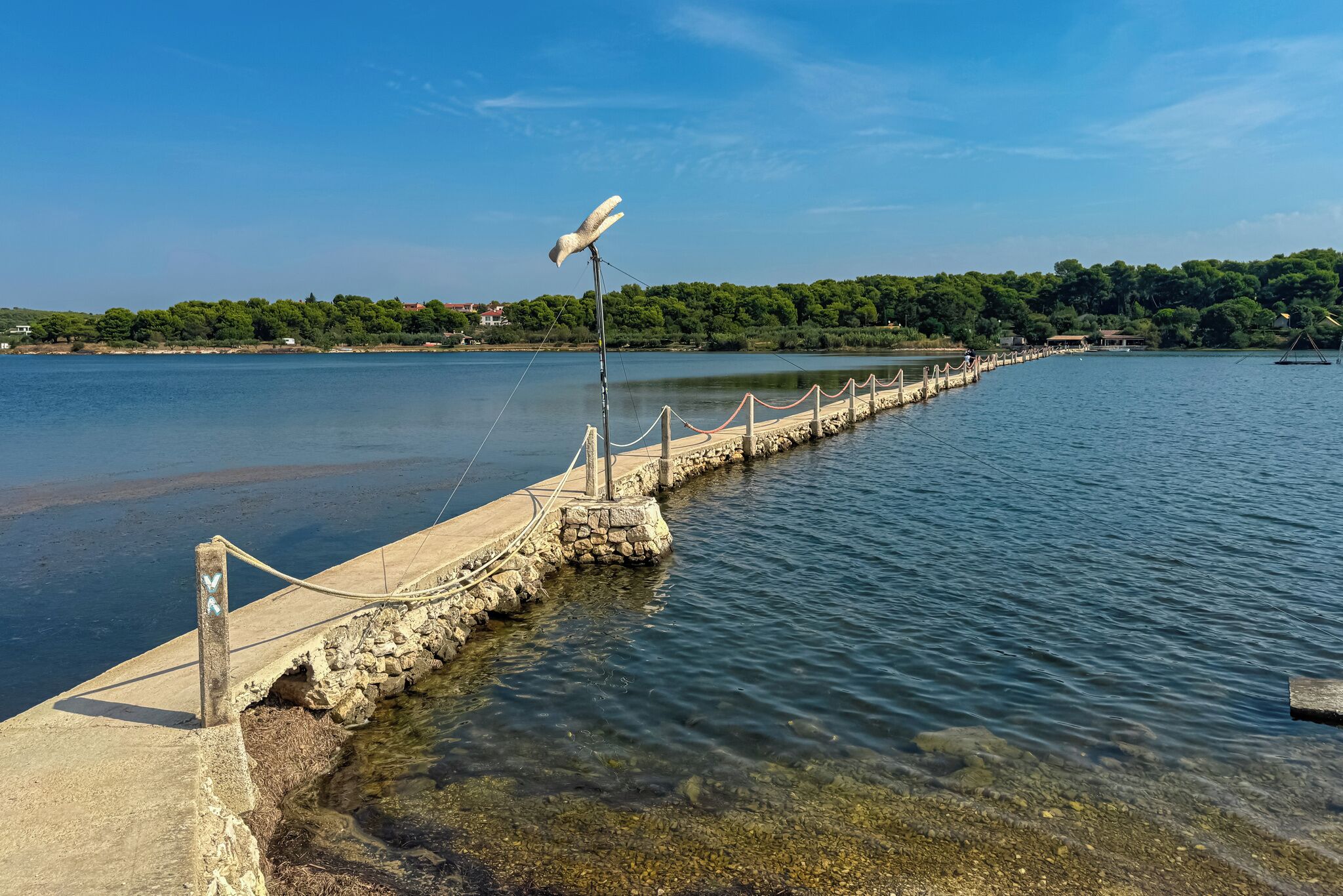 Een steiger die zich uitstrekt over een rustig meer, met een standbeeld van een vogel op een paal, omgeven door helder water en een verre oever begroeid met bomen.