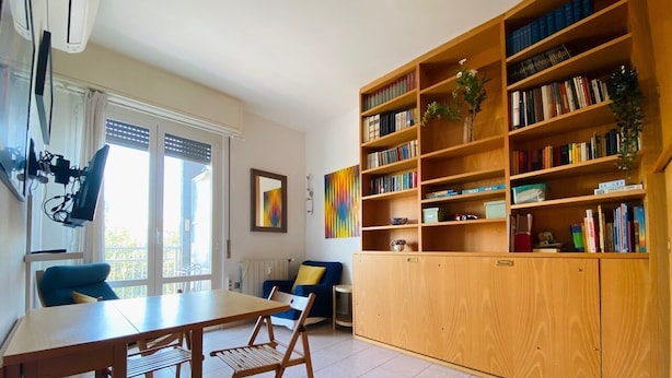 A room with a wooden table and chairs, a bookshelf, and a window with curtains. - Fornari 48 Comfortable Flat (Milan)
