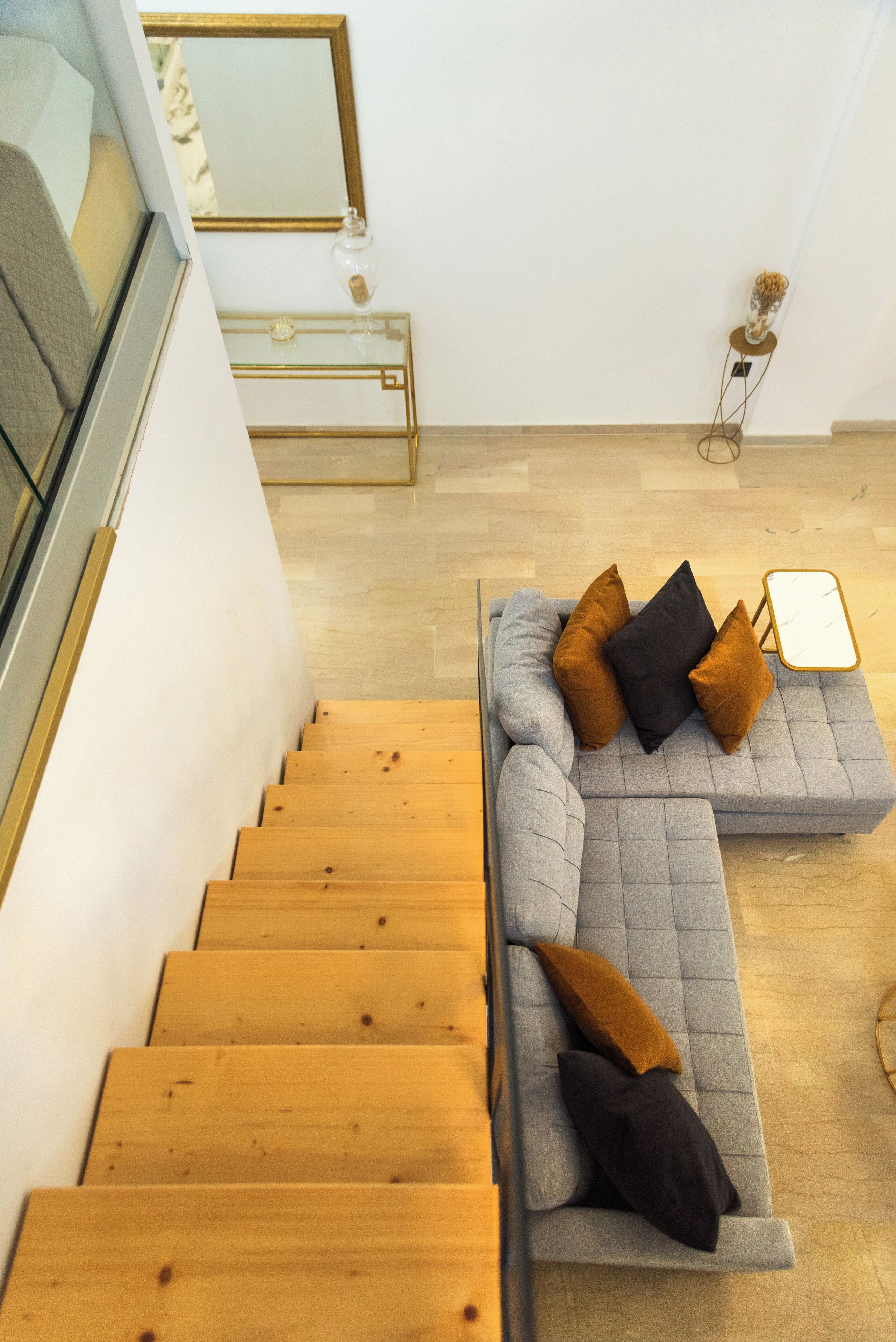 A modern living room with a grey sectional sofa, wooden stairs, a glass coffee table, and a mirror on the wall.