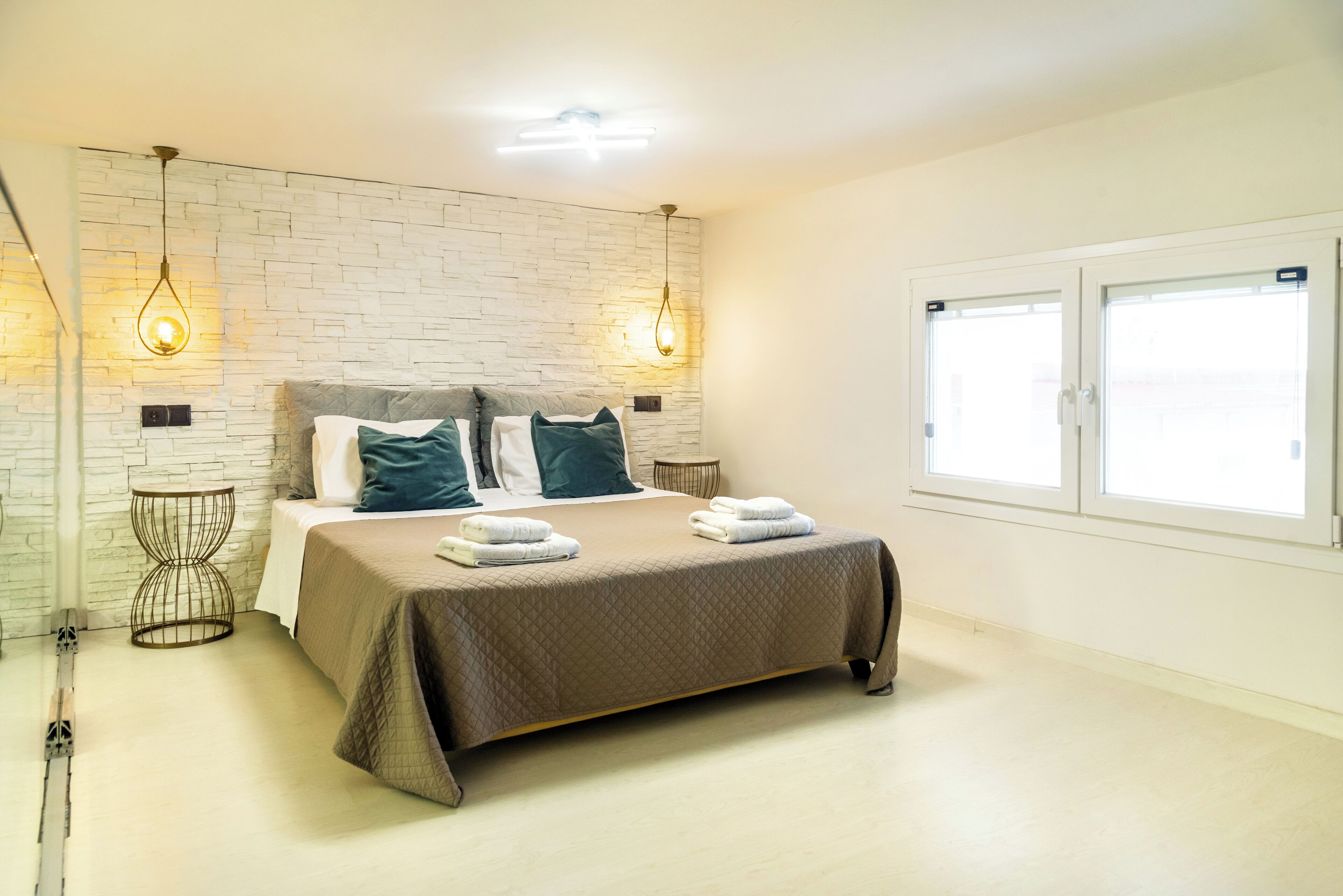 Quarto moderno com parede de tijolos, cama grande com cobertura acolchoada, duas luminárias de cabeceira e janela com persianas.