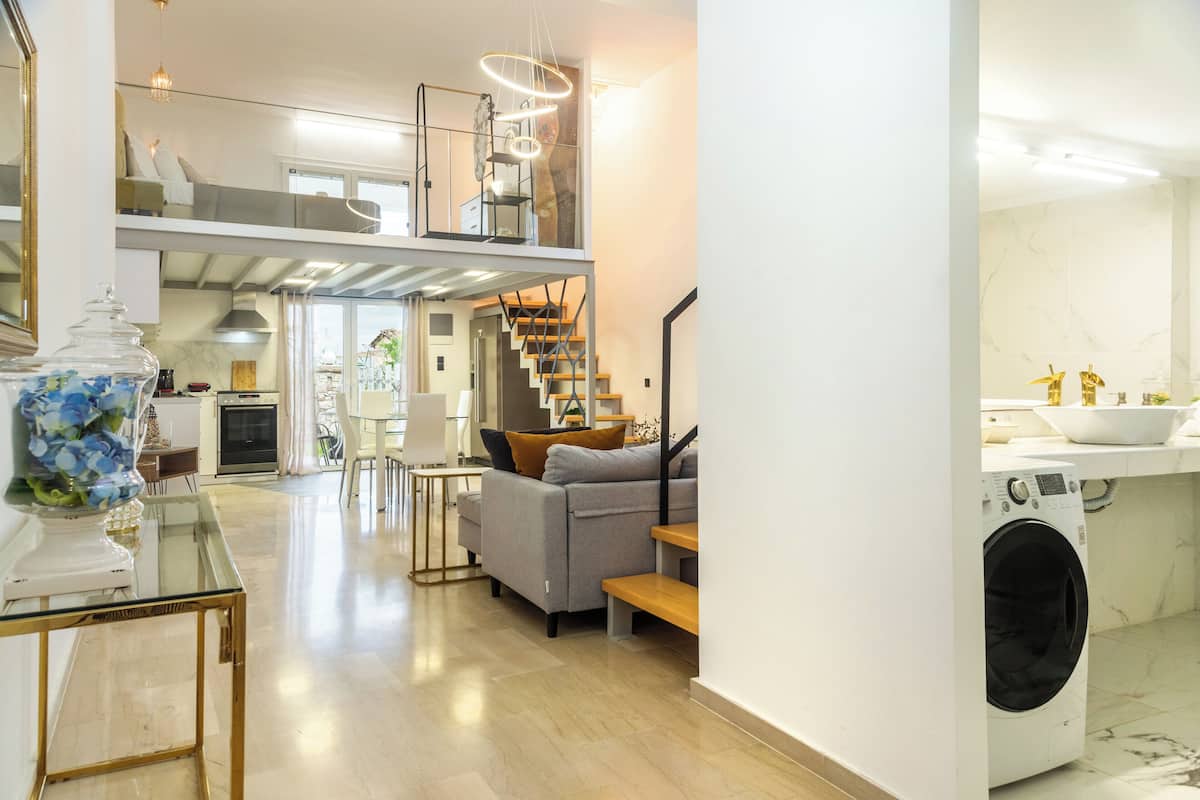 A modern kitchen with a built-in washer and dryer, a dining area with a glass table, and a staircase leading to a loft bedroom.