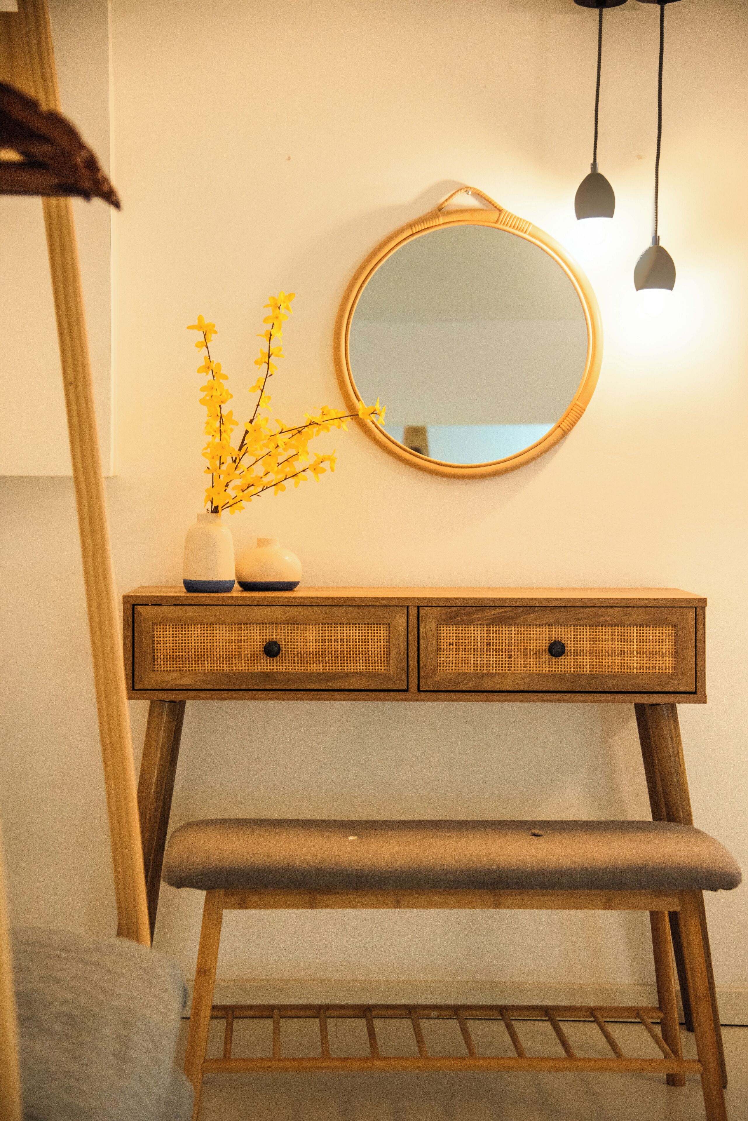 Een houten wastafel met een ronde spiegel, twee kleine vazen met gele bloemen en een bankje.