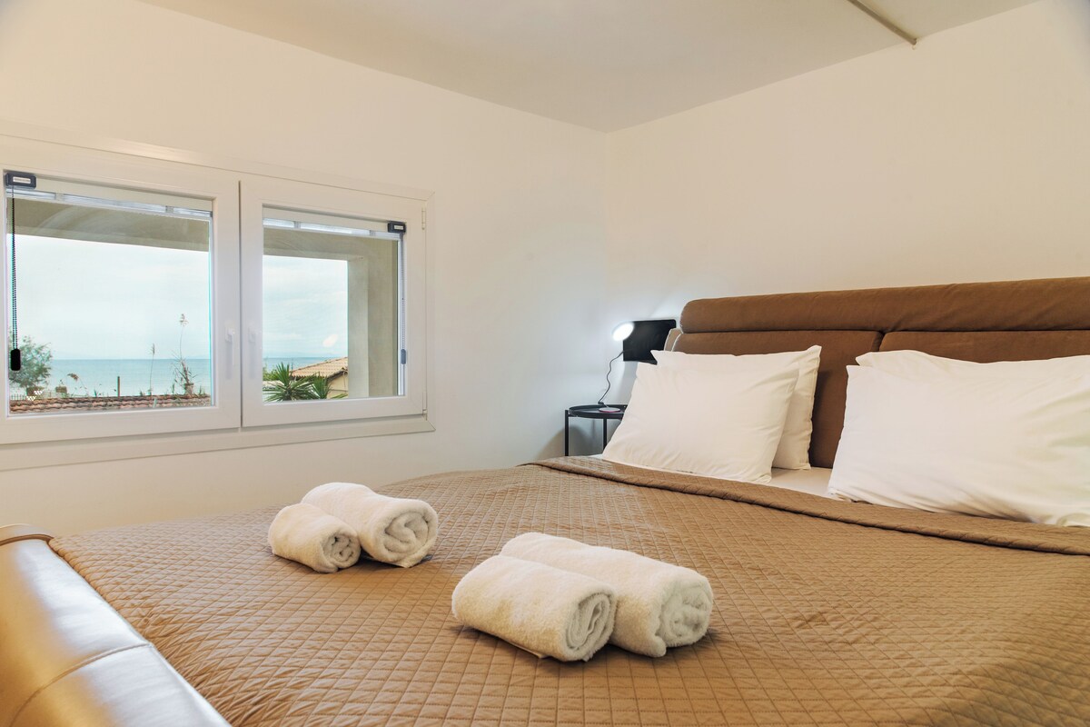 A bedroom with a bed, two windows, a nightstand, and a view of the beach.