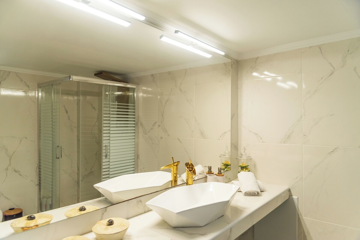 A modern bathroom with a large mirror, two sinks, a shower, and a marble backsplash.
