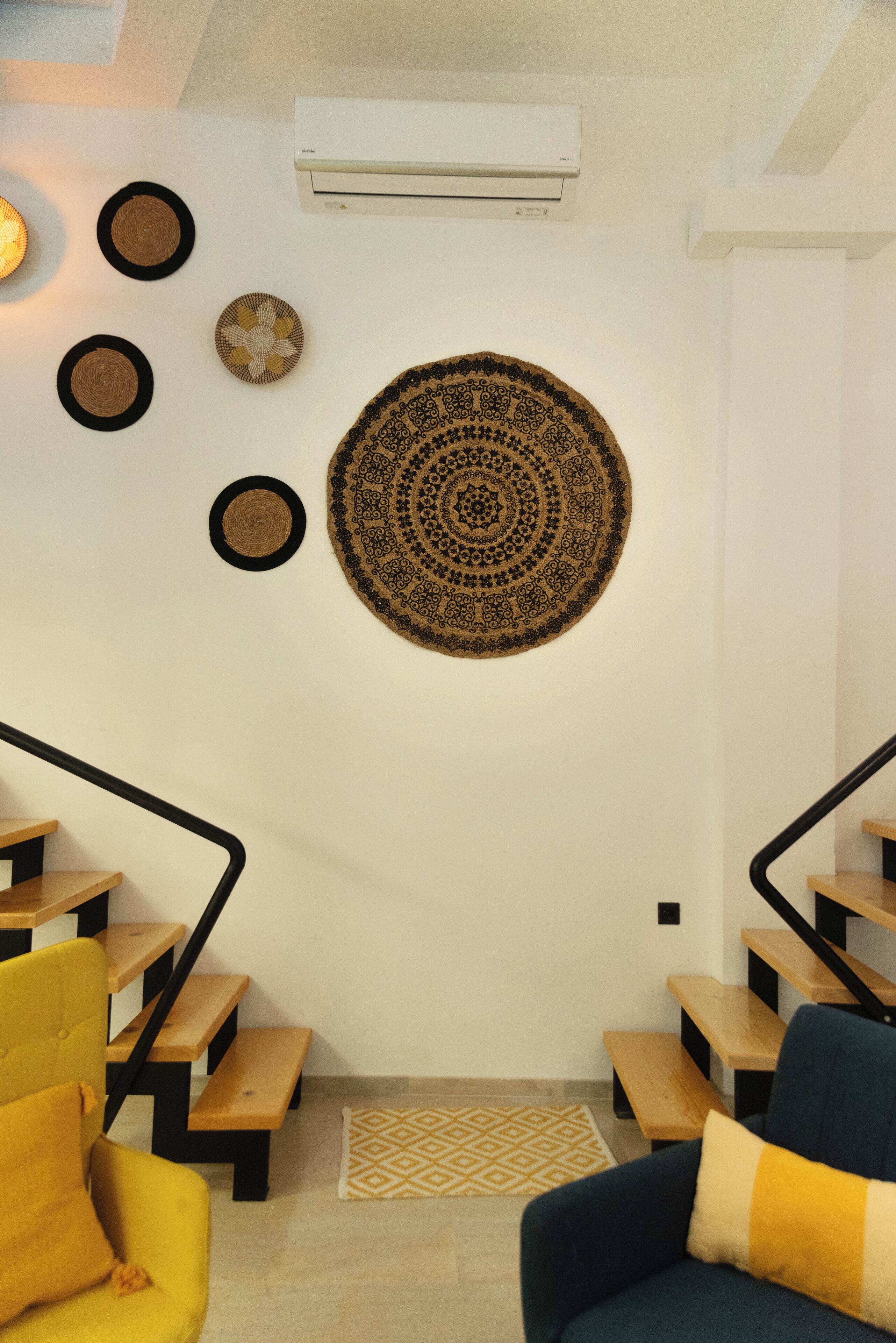 A modern living room with a staircase, a large circular wall hanging, and a ceiling-mounted air conditioner.