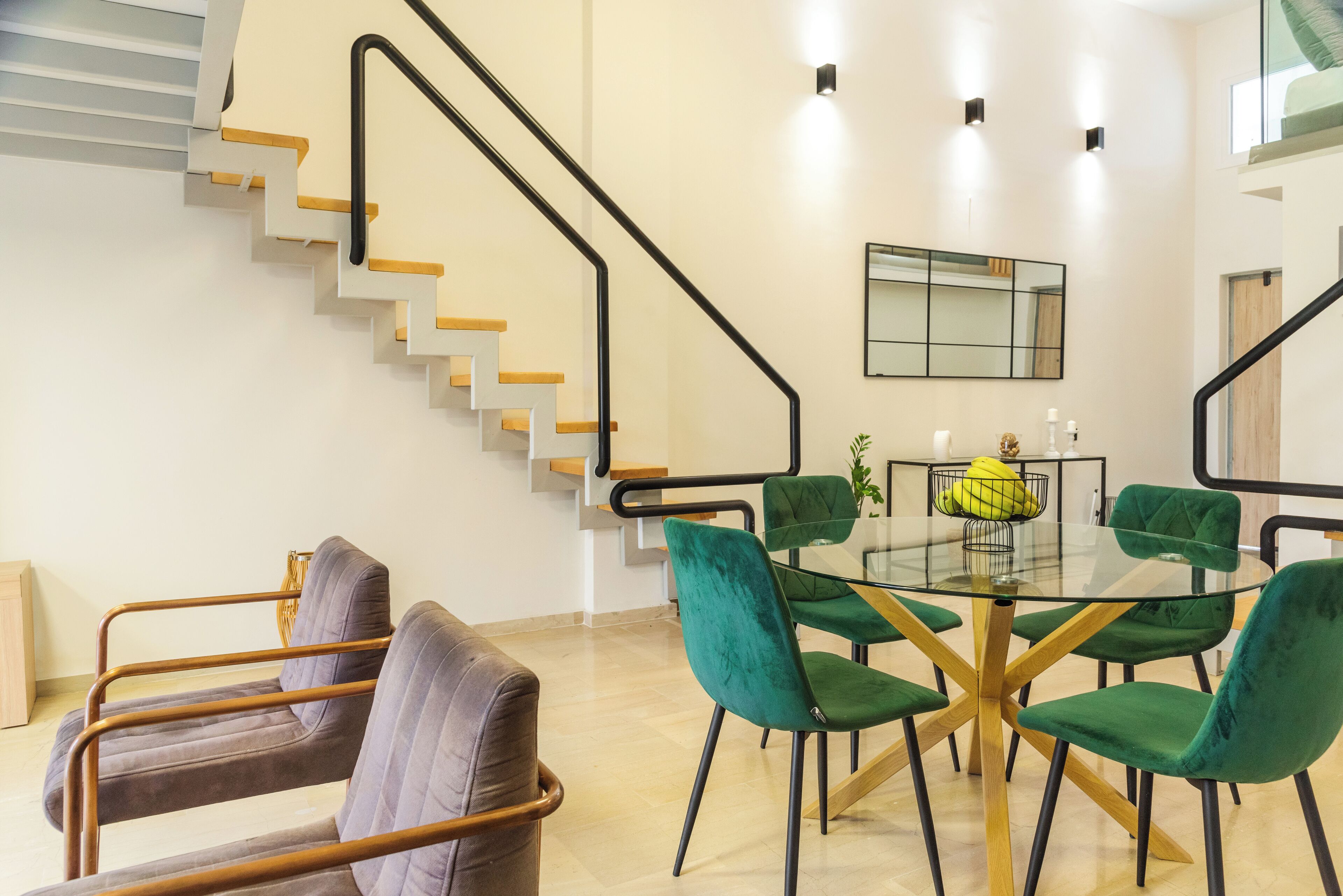 A modern living room with a glass dining table surrounded by green chairs, a staircase with wooden steps, and a minimalist design.