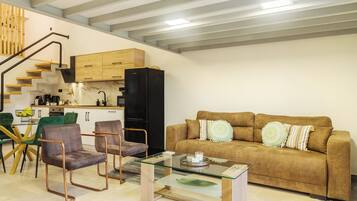 Una sala de estar moderna con un sofá marrón, dos sillones, una mesa de centro de vidrio y una zona de cocina con armarios de madera y un refrigerador negro.
