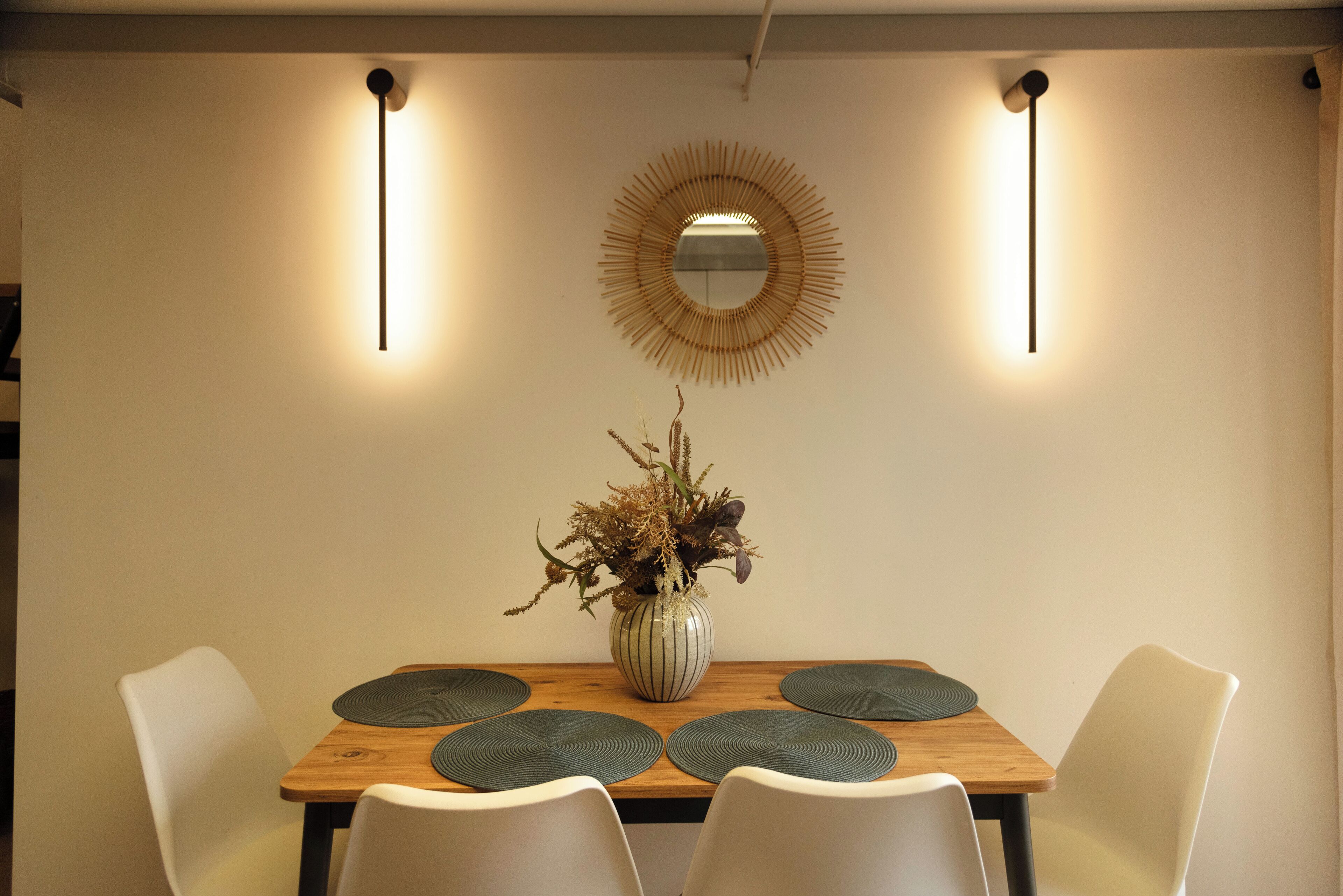 Uma sala de jantar com mesa de madeira, cadeiras brancas, um vaso com plantas secas e um espelho redondo na parede.